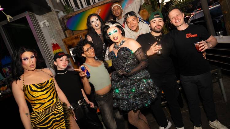 Three drag queens pose with a tour group outside of a club with different pride flags on display behind them.