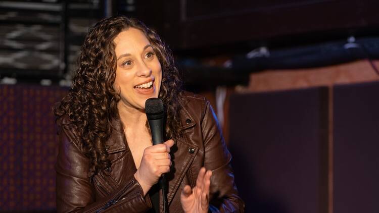 Jenny Hagel talks into a microphone in a dimly lit comedy club.