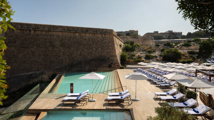 Outdoors at The Phoenicia Malta, showing rows of sun loungers surrounded by a backdrop of ancient buildings 