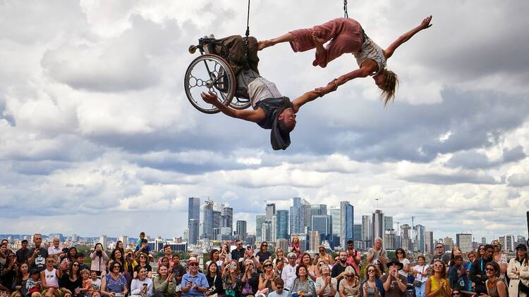 Two aerial performers, one in a wheelchair, hold hands upside down in front of a crowd of spectators, with the buildings of Canary Wharf in the background at Greenwich Fair in 2023