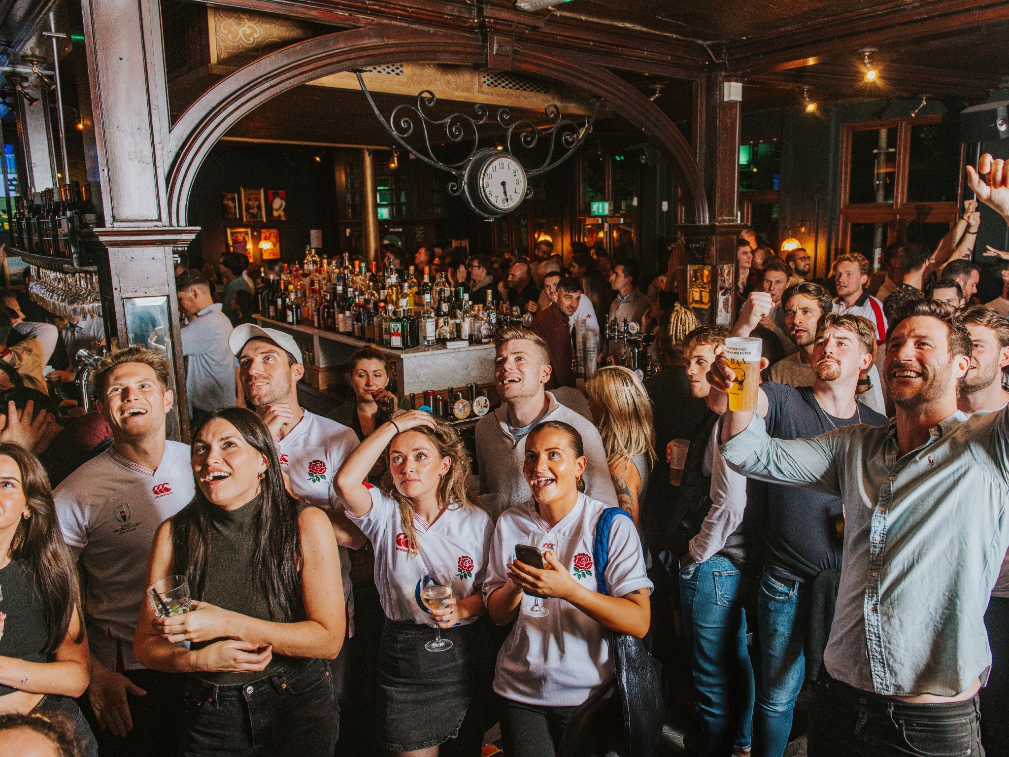 A large crowd of rugby fans, some wearing England shirts, standing in a traditional pub staring at a screen