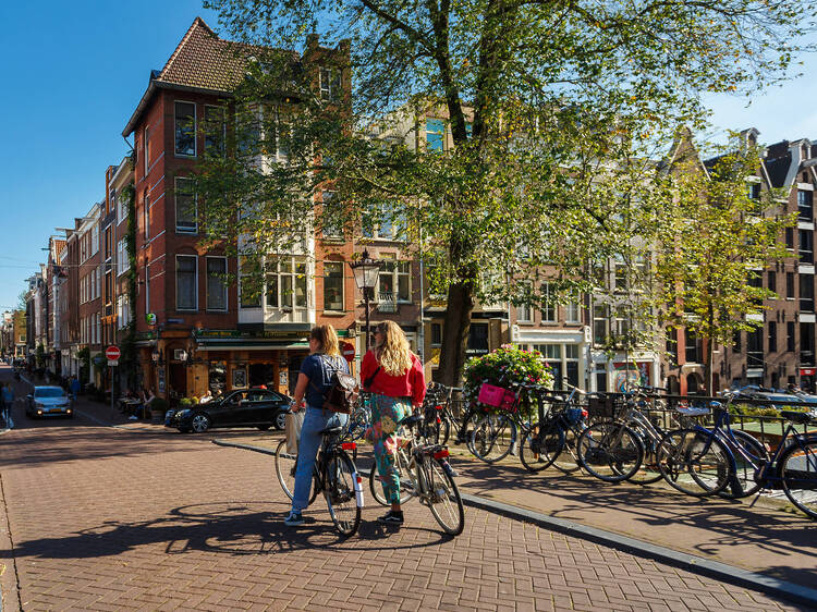 Two young woman with bikes are walking on the bridge of Amsterdam at sunny day