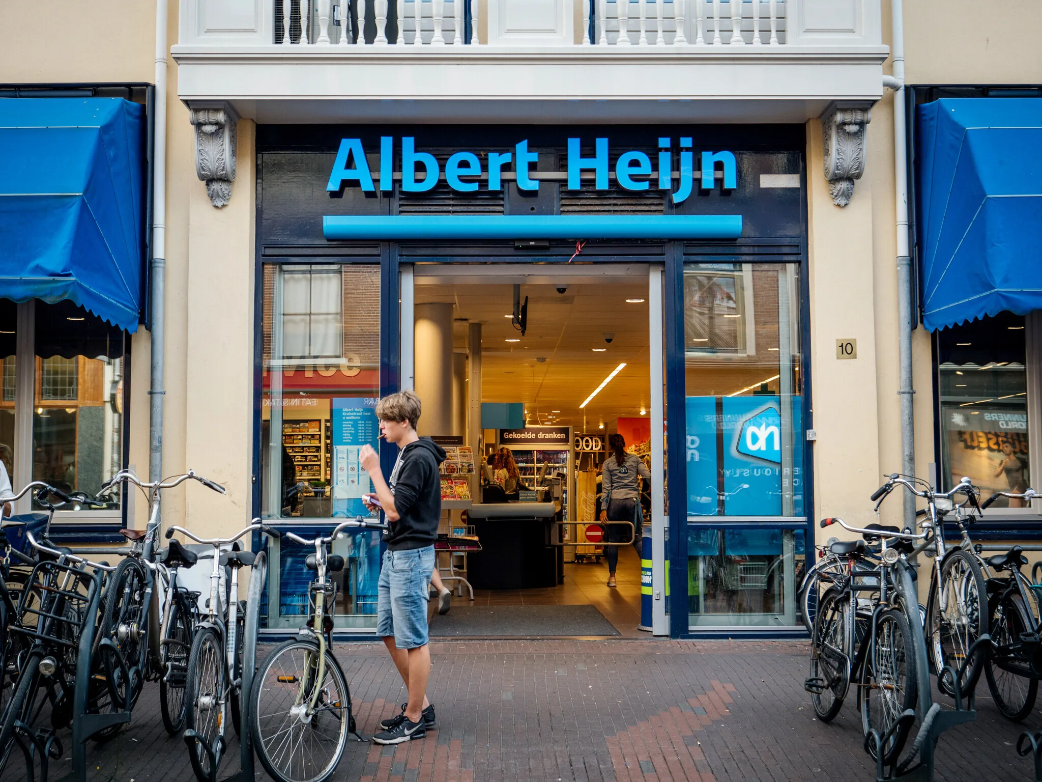 Front view of Albert Heijn Dutch supermarket chain, founded in 1887 in Oostzaan, Netherlands on Kruisstraat 10 street with young man eating a chocolate outside