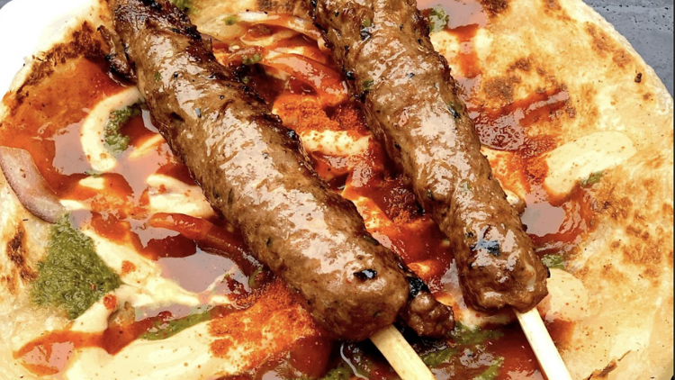 Two grilled meat kebabs on skewers served over a flaky flatbread with swirls of creamy sauces, green chutney, and red chili oil.
