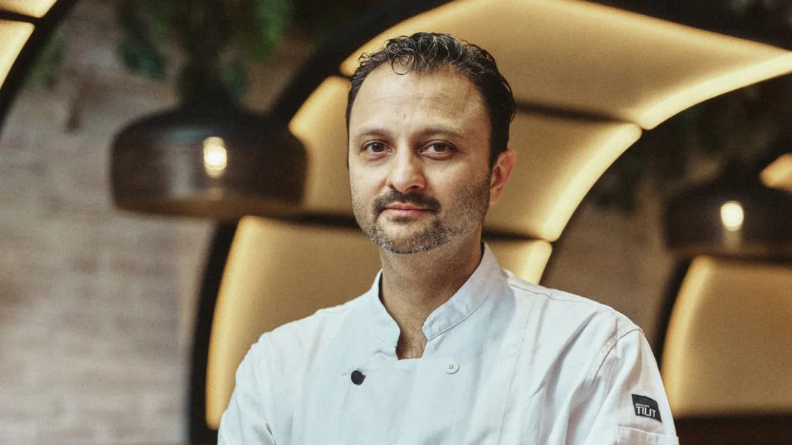Chef Chintan in a white chef’s coat stands with his arms crossed inside a warmly lit restaurant with yellow awnings in the background.