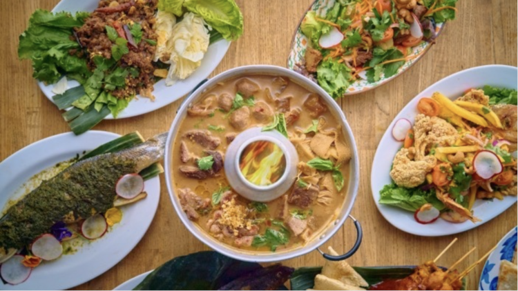 A colorful spread of Thai dishes from Kam Rai Thai, including a whole grilled fish topped with herbs, a fiery hot pot filled with meat and vegetables, and assorted salads with fresh greens, radishes, and crispy toppings.