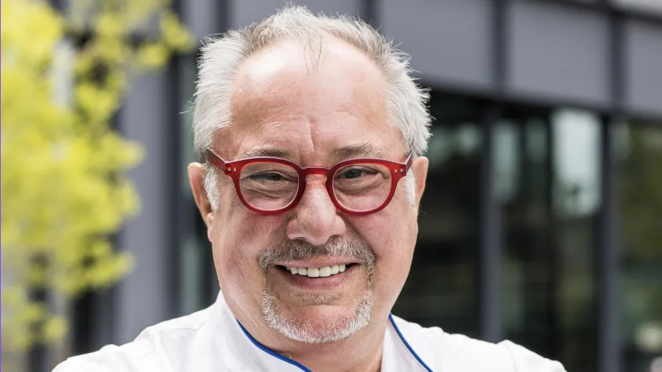 Chef Michael Ayoub of Fornino, wearing red glasses and a white chef’s coat, smiling outdoors in front of a modern building.