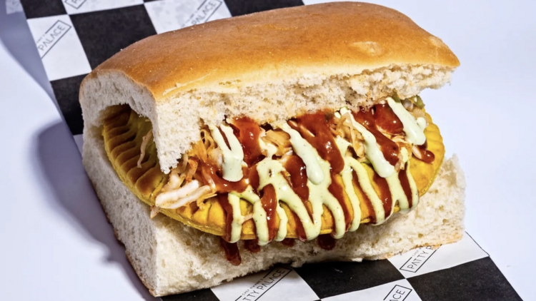 A Jamaican patty sandwich from Patty Palace, served inside soft white bread and topped with shredded cabbage, red sauce, and green crema drizzle.