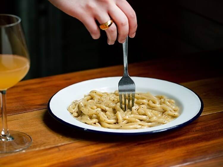 Pici pasta in white plate with glass of orange wine
