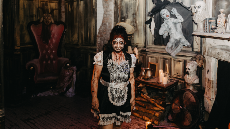 A woman dressed as a haunted maid, covered in facepaint and blood, standing in a scary haunted room