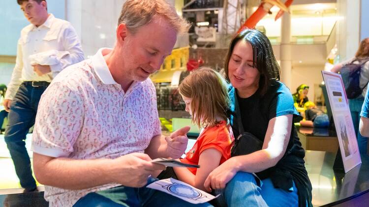 Two parents and a child solve a puzzle together at an "Escape the Museum" event. Two parents and a child solve a puzzle together at an "Escape the Museum" event.