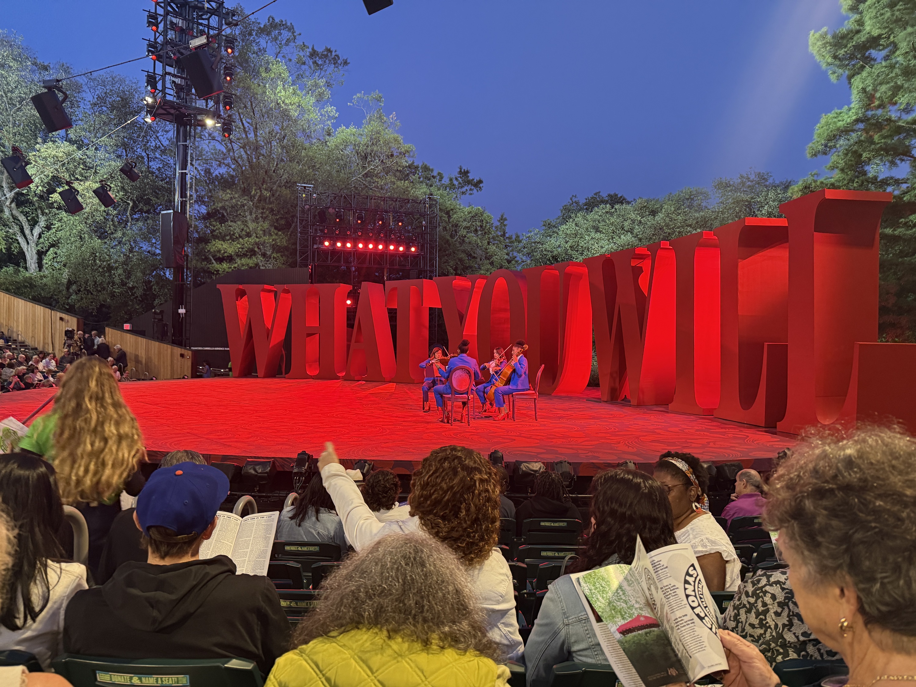 Picture of the Delacorte Theater stage with big letters reading 'WHAT YOU WILL' in red lighting. Musicians sit on the stage in purple suits.