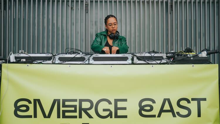 Emerge East free festival A DJ playing music from a desk covered with a sign saying 'Emerge East'