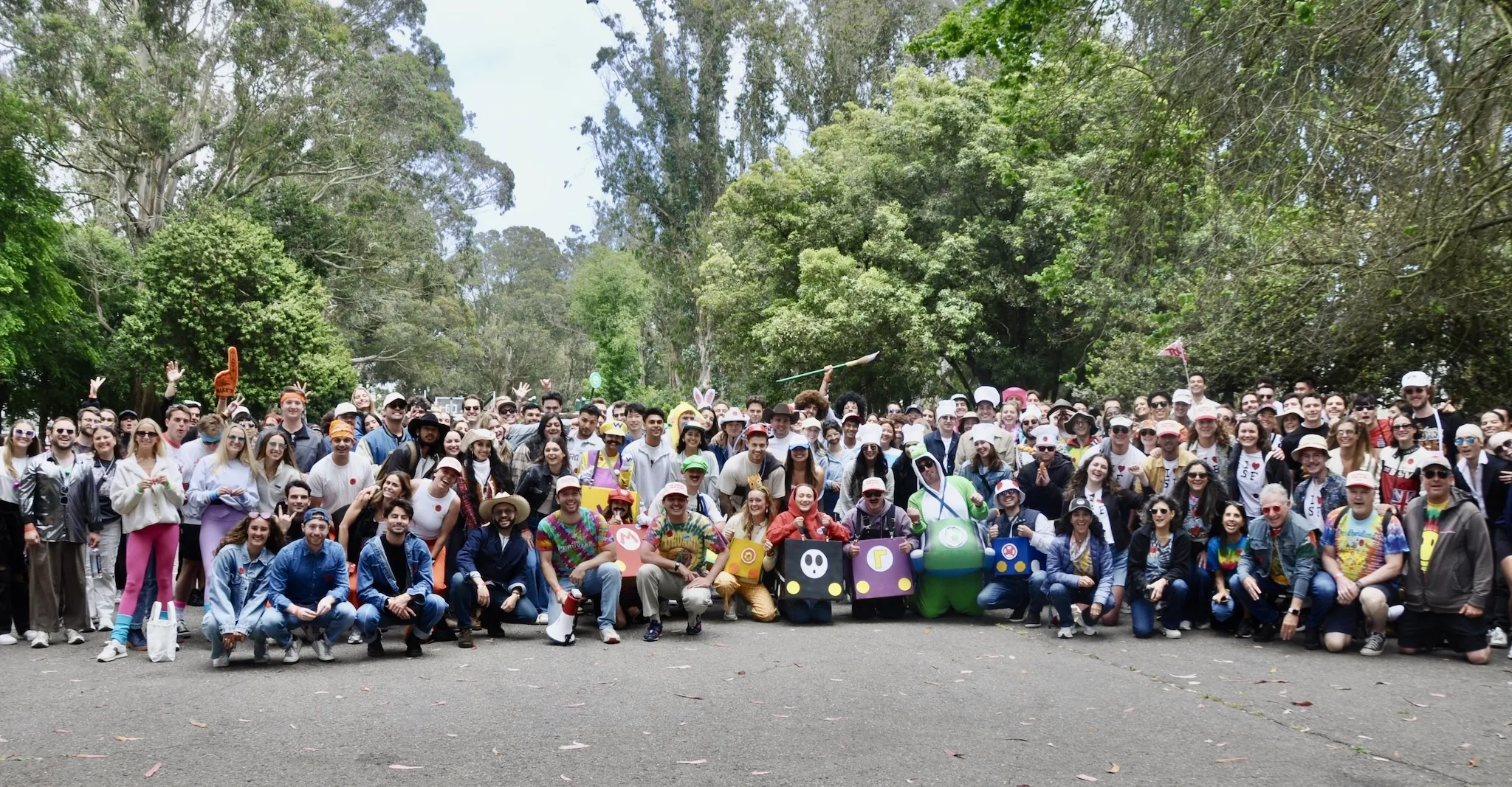 A large group of people in costume pose in front of a thicket of trees.