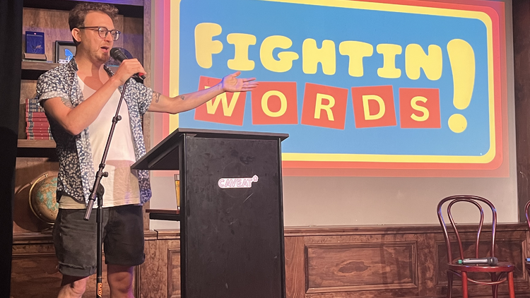 A man onstage at a podium with a microphone infront of a projection that says "FIGHTIN WORDS!"