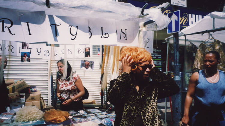 Photograph of a Black woman wearing a blonde wig at a market in England 