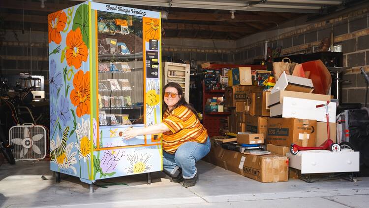 Steph Krim, founder of Good Things Vending, embraces a colorfully painted Good Things Vending machine in a garage piled with boxes.