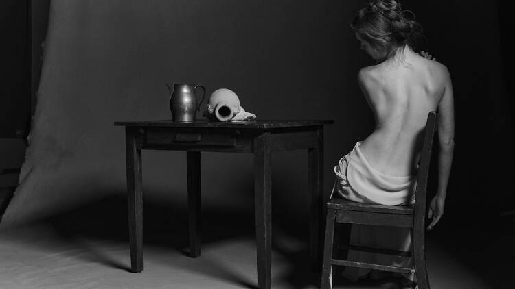 A topless woman sits with her back to the camera next to a table with a skull and pitcher on it.