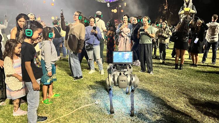 A robot dog surrounded by audience members in headphones in The Cortège.