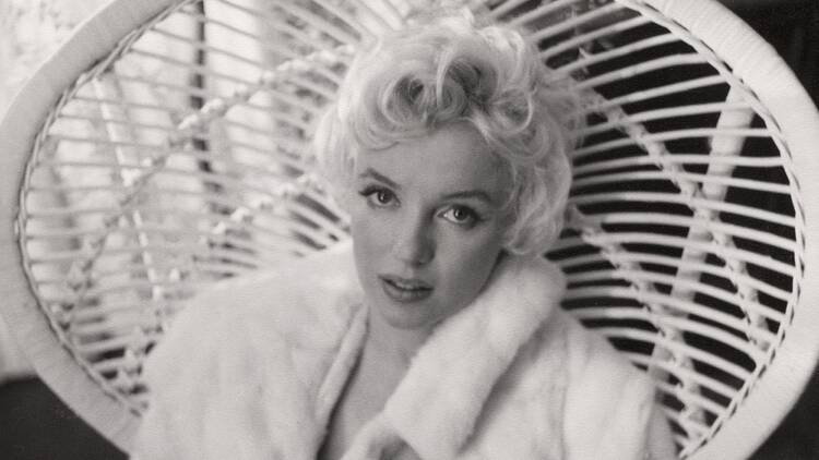 A black and white photograph of Marilyn Monroe wearing a fur coat and staring at the camera while reclining on a wicker structure