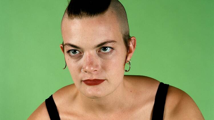 A woman with a wide black and red mohawk wearing a black tank top poses in front of a green background