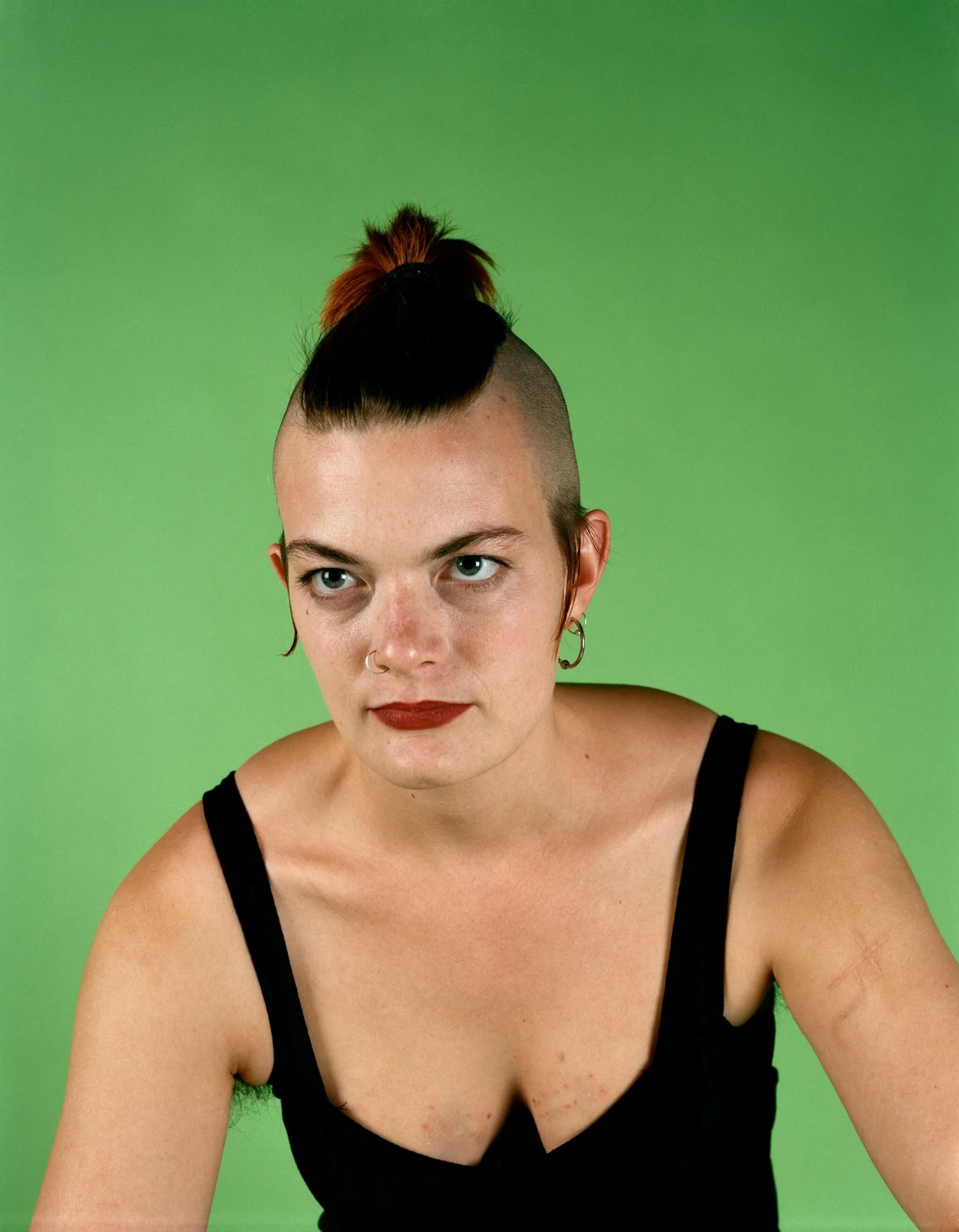 A woman with a wide black and red mohawk wearing a black tank top poses in front of a green background