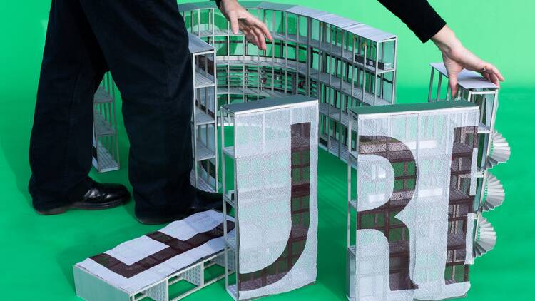 A faceless person rearranges pieces of a small-scale architectural model. 