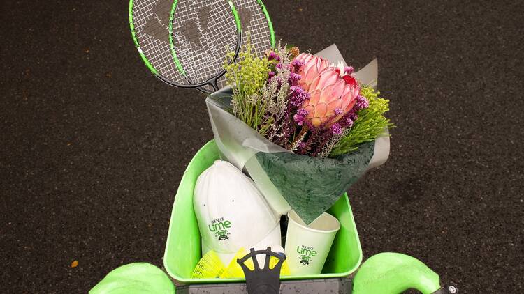House of Lime Lime bike basked carrying a bunch of flowers, a paper cup, two badminton rackets and a white baseball cap