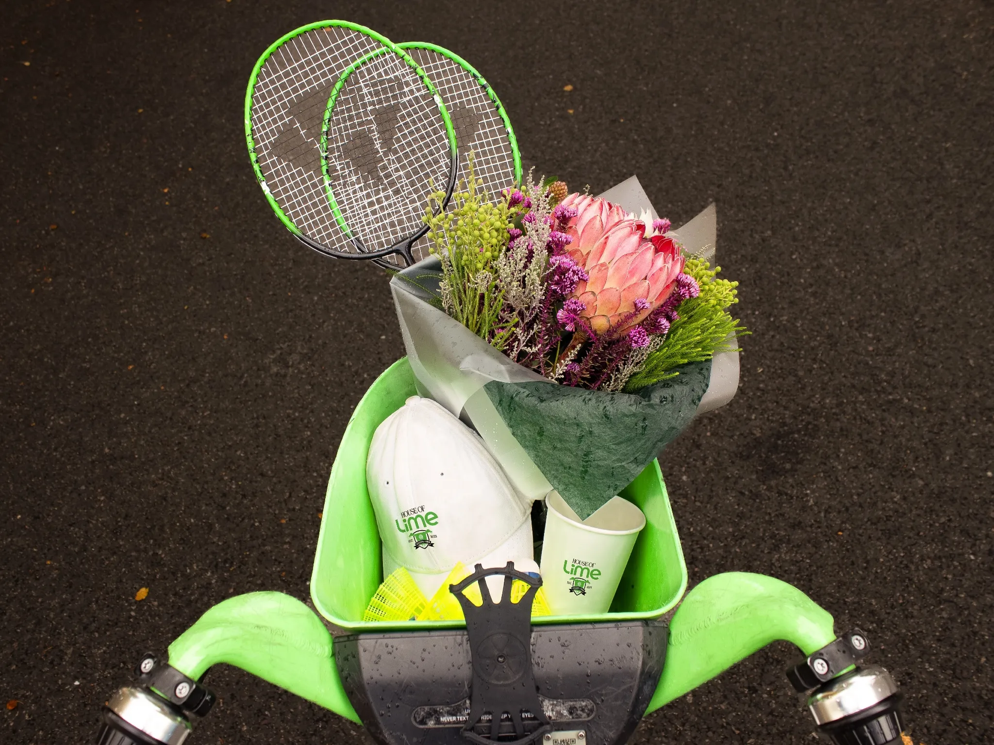 Lime bike basked carrying a bunch of flowers, a paper cup, two badminton rackets and a white baseball cap
