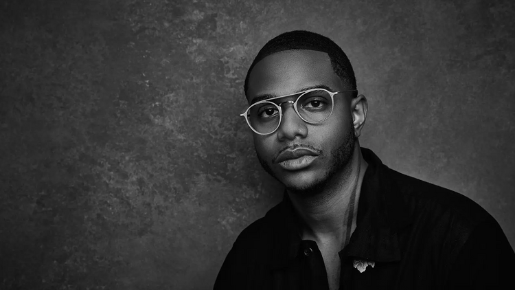 Chef Kwame Onwuachi pictured in black and white against a textured background. He wears a dark shirt and ligth rimmed glasses. 