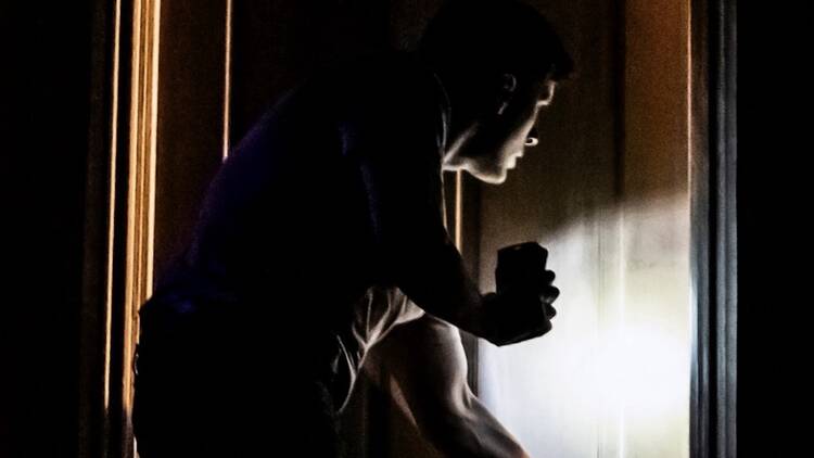 A dimly lit person peers past a door with their cell phone flashlight