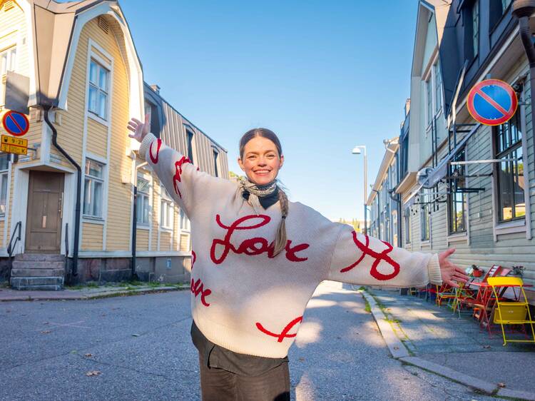 Hymyilevä nainen poseeraa puutalojen keskellä paidassa jossa lukee Love