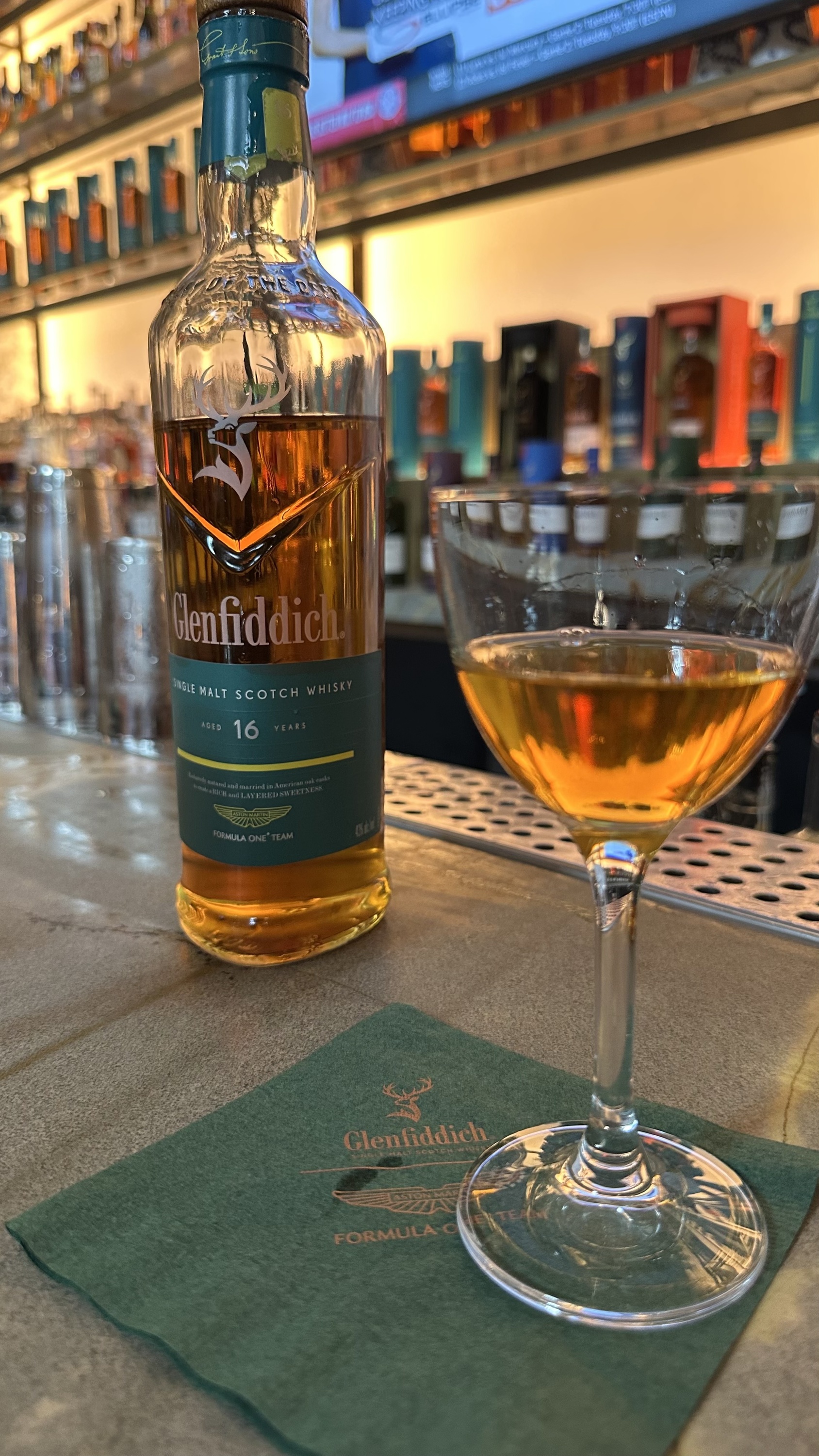 A bottle of whisky sits next to a glass of whisky at a bar.