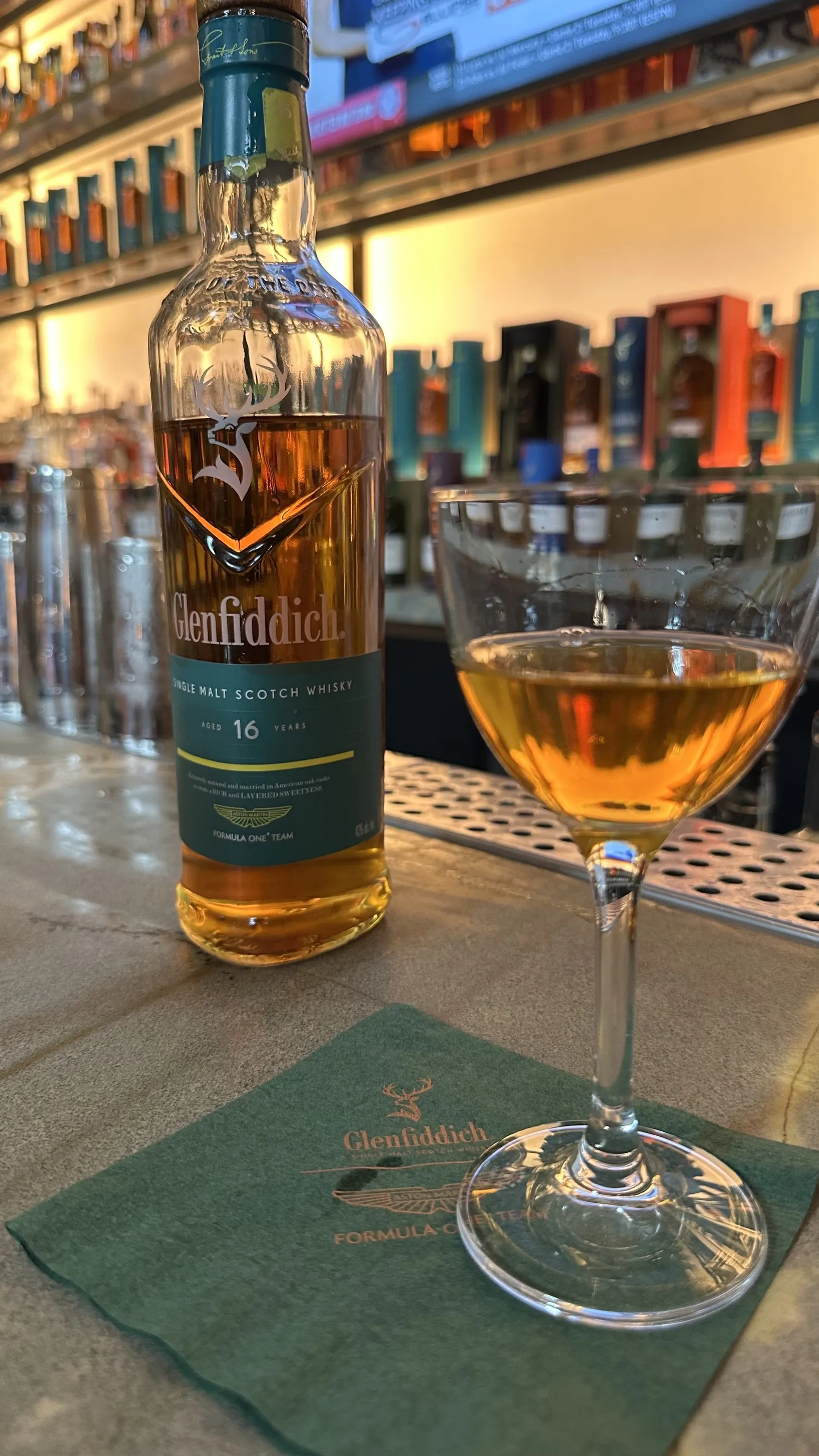 A bottle of whisky sits next to a glass of whisky at a bar.