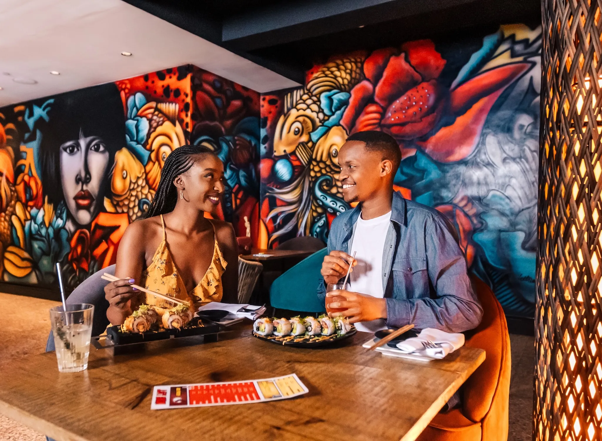 Two people sitting a table over Japanese food with colorful murals behind them