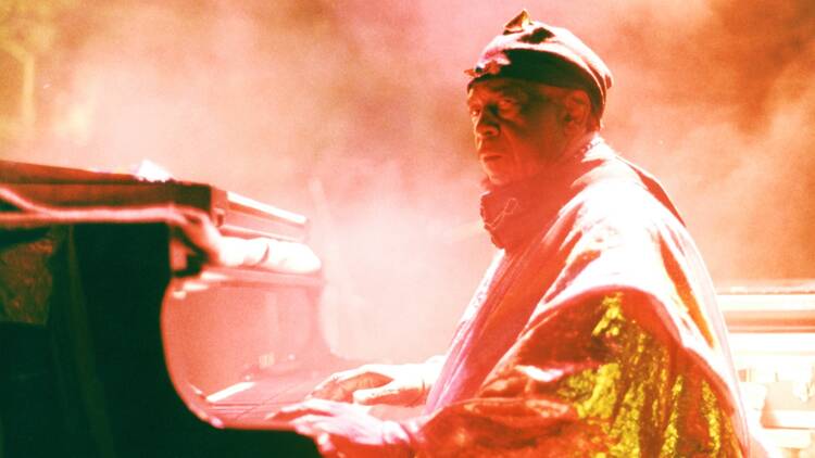 Sun Ra in orange light and haze plays piano and glares at the camera. Sun Ra in orange light and haze plays piano and glares at the camera.