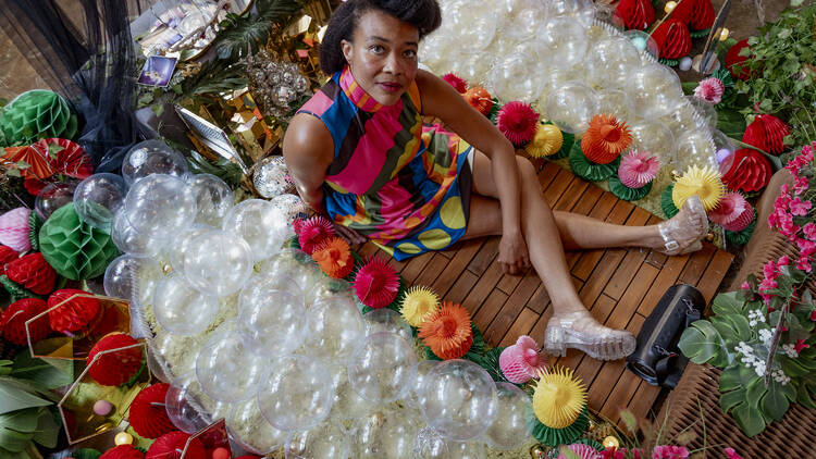 A Black woman in a colorful dress sits on the ground amid a colorful art exhibit.