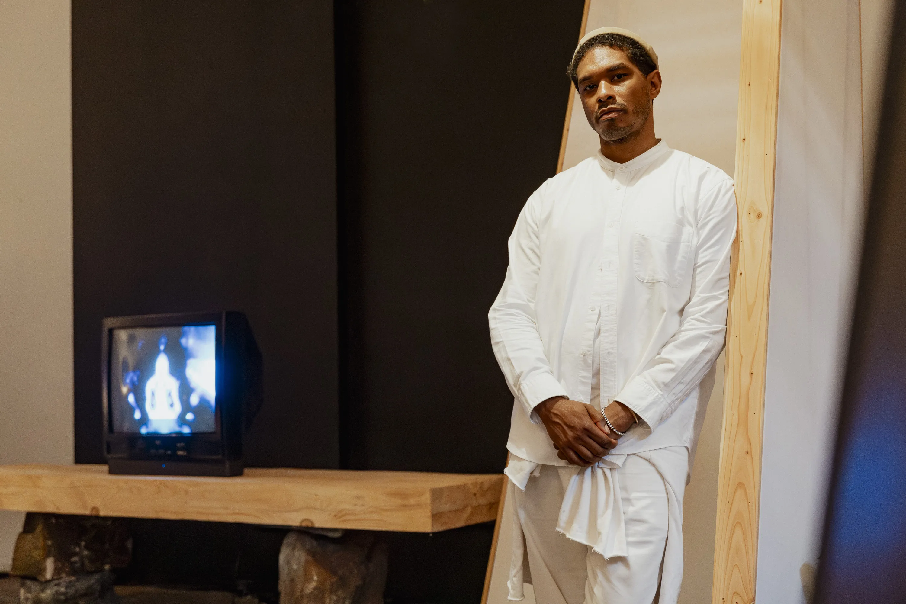 An Black male artist in a white outfit stands next to his work, an altar with a tube TV.