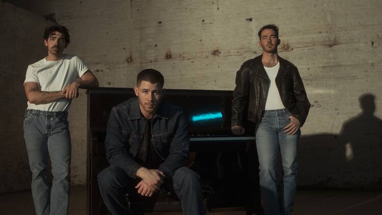 The Jonas Brothers pictured standing around a piano in a warehouse. The Jonas Brothers pictured standing around a piano in a warehouse.