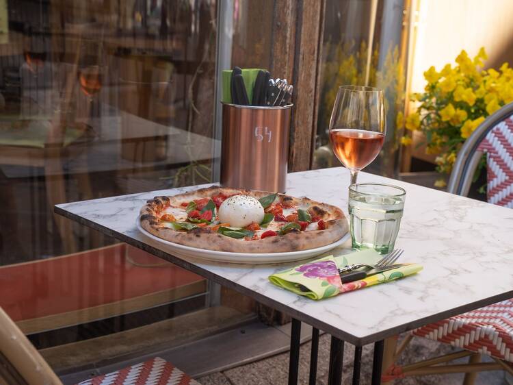 An outdoor table is set with a burrata pizza topped with basil and cherry tomatoes, accompanied by rosé wine and water. An outdoor table is set with a burrata pizza topped with basil and cherry tomatoes, accompanied by rosé wine and water.