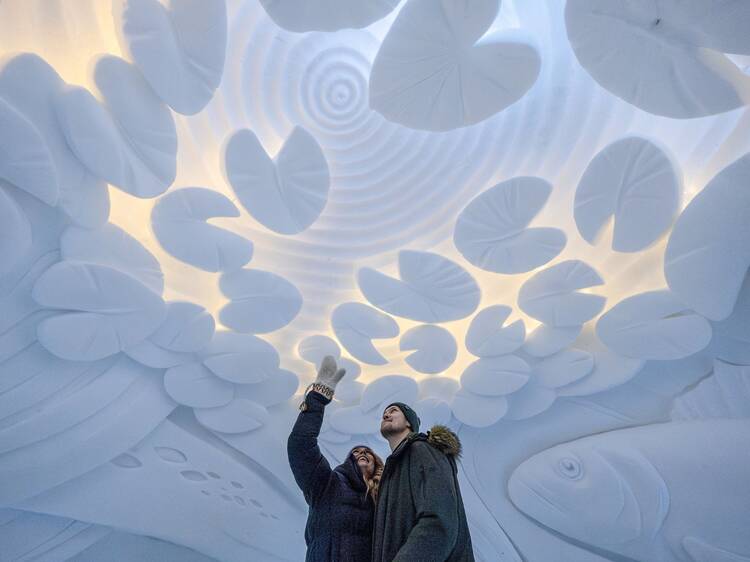Two people stand beneath a stunning circular ceiling installation made of white, leaf-like structures illuminated by soft lighting. Two people stand beneath a stunning circular ceiling installation made of white, leaf-like structures illuminated by soft lighting.