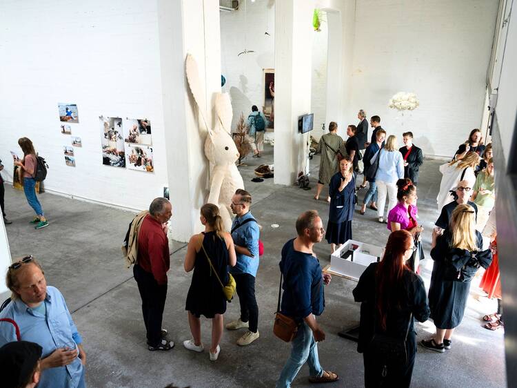 A bright gallery space with visitors engaging around wall-mounted photographs, a central rabbit sculpture, and various contemporary installations.