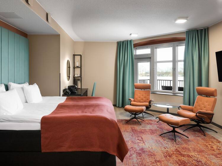 A stylish hotel room featuring a large bed, orange lounge chairs, teal curtains, and a sleek desk area in a modern setting. A stylish hotel room featuring a large bed, orange lounge chairs, teal curtains, and a sleek desk area in a modern setting.