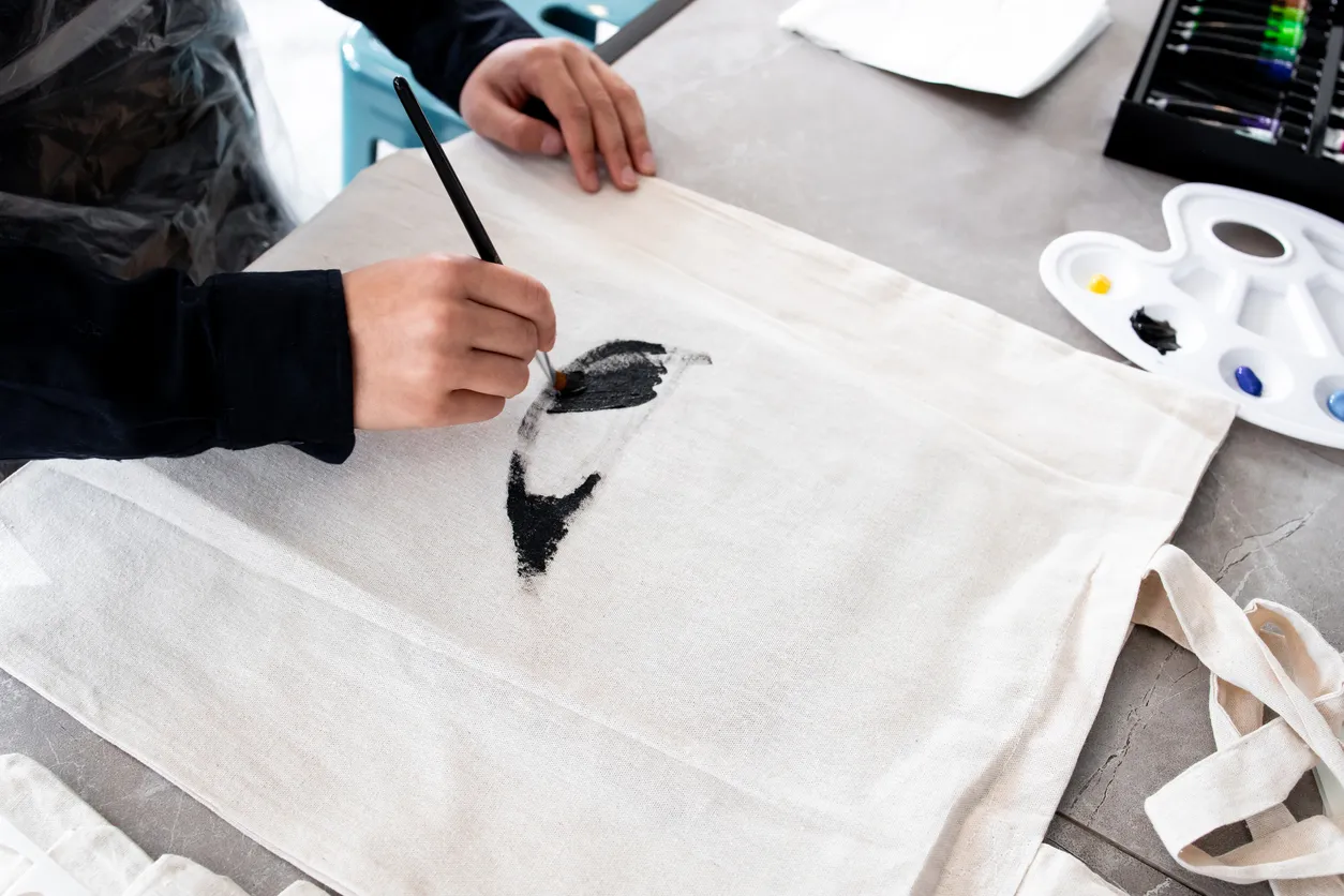 Person painting design on canvas bag with black brush strokes in art studio.