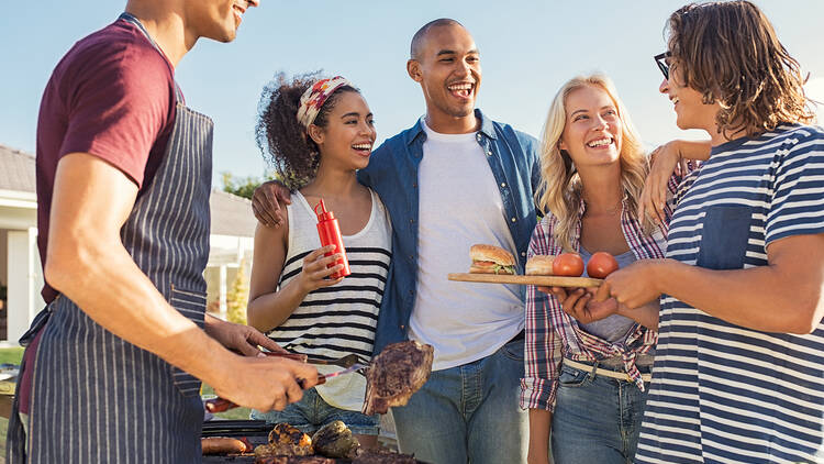 Friends enjoying lunch barbeque/braai Young multiethnic friends having fun grilling meat enjoying bbq party. Group of happy guys and girls cooking and eating at barbecue dinner outdoor. Men and women standing around grill, chatting, drinking and eating.