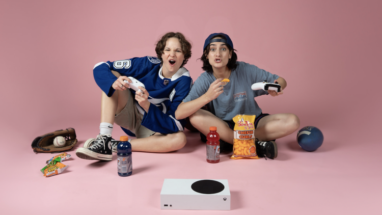 Two girls sitting on a pink background with snacks and video games