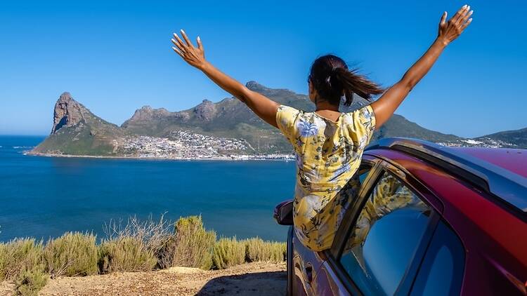 woman outside car window road trip with hands up, car at Chapmans Peak Drive in Cape Town South Africa looking out over ocean 1407971883