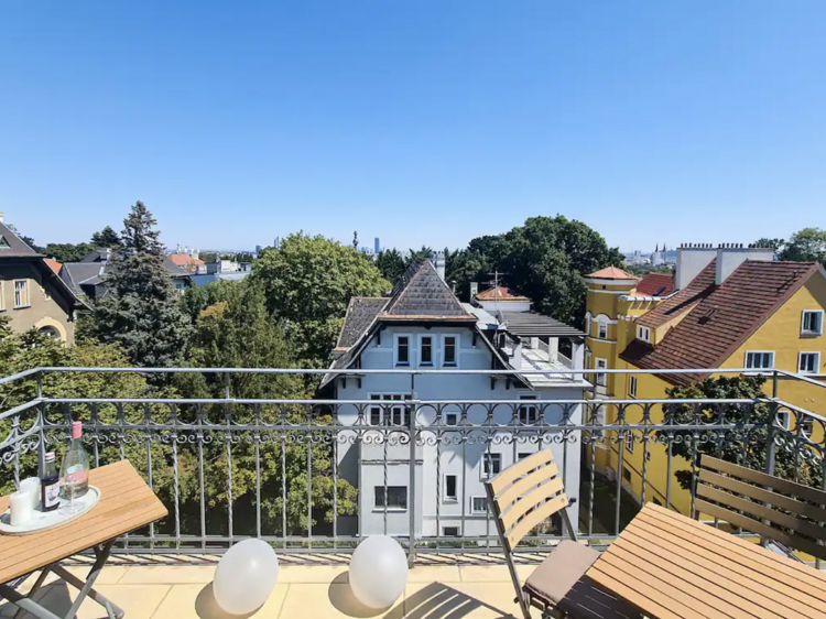 Photo of a sun drenched balcony overlooking a bright blue house and a bright yellow house and lots of green trees 