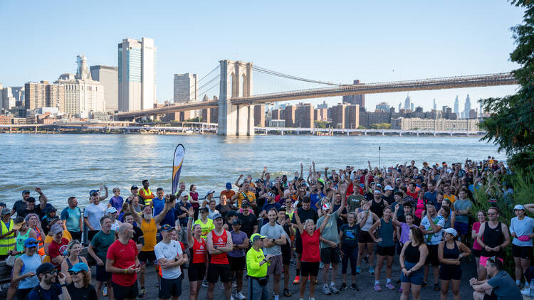A large group of people gathered in the brooklyn bridge park, in front of the brooklyn bridge, preparing to run a 5k A large group of people gathered in the brooklyn bridge park, in front of the brooklyn bridge, preparing to run a 5k