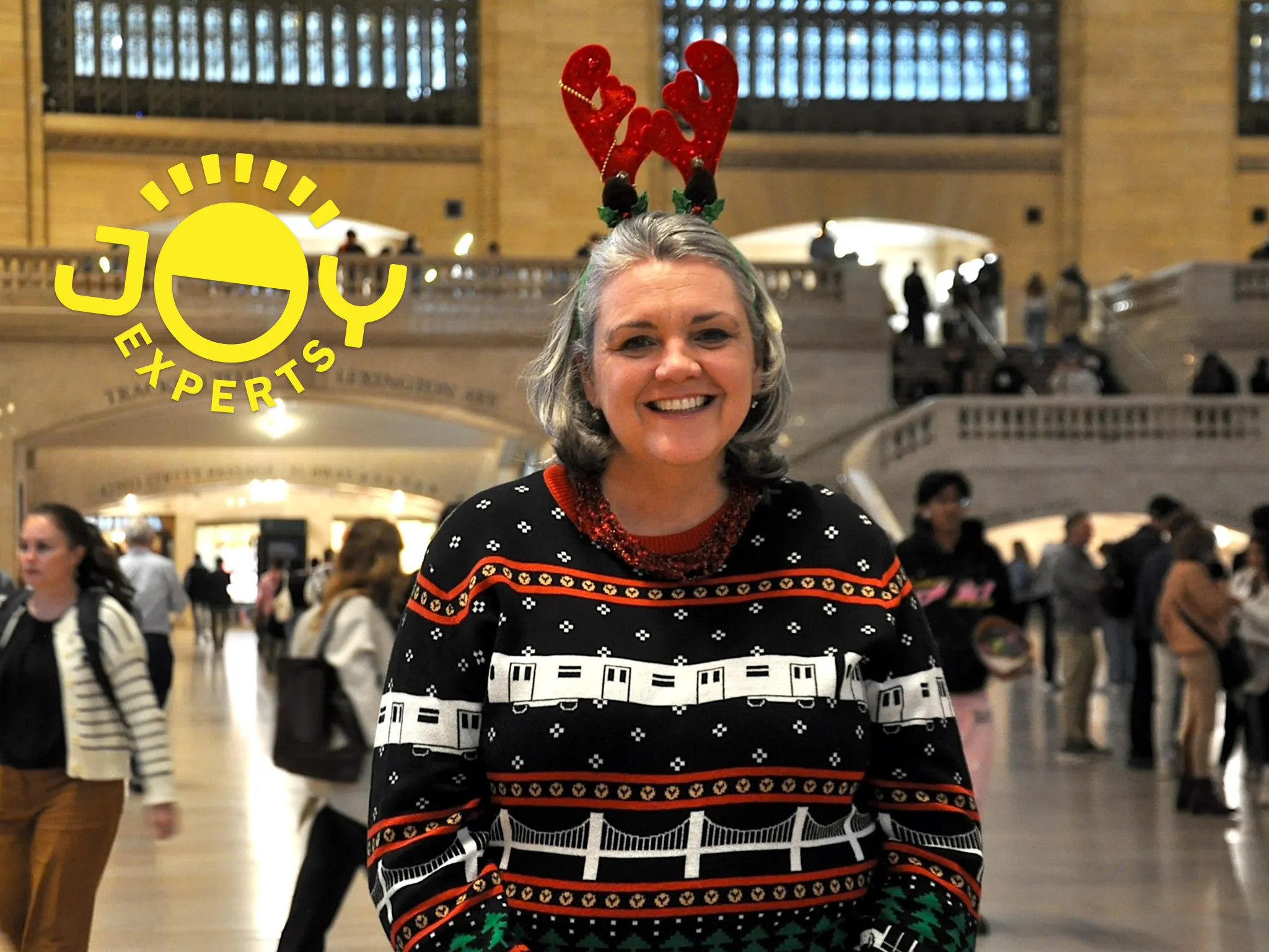 A woman wearing a holiday train sweater and reindeer antlers poses in Grand Central.